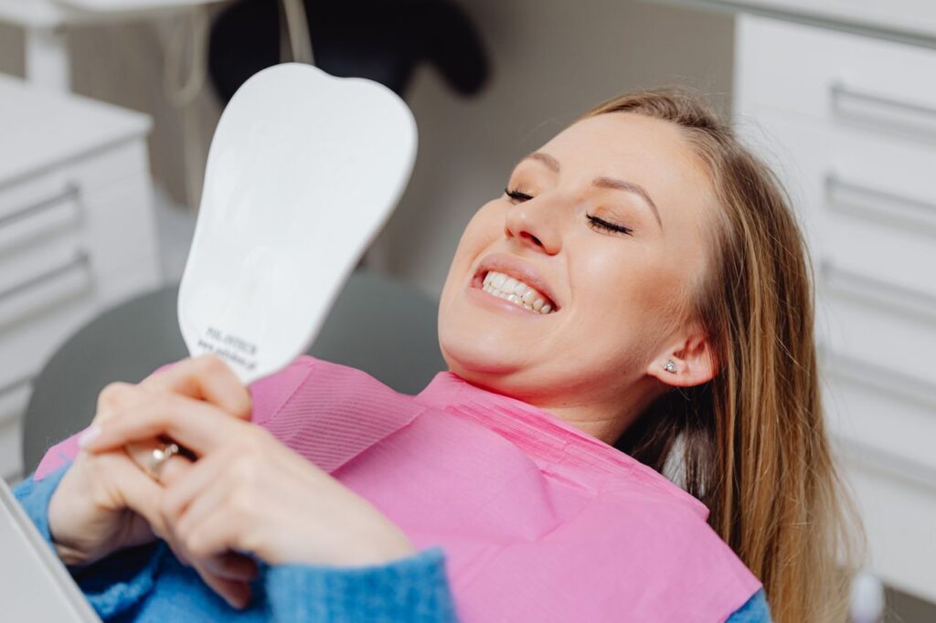 pexels-photo-6627574-6627574 A woman at a dental clinic smiles while holding a mirror, showcasing her dental treatment results.